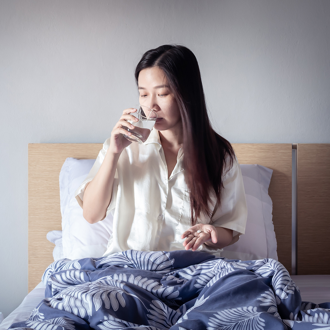 A young woman takes a pill with a glass of water from her bed.