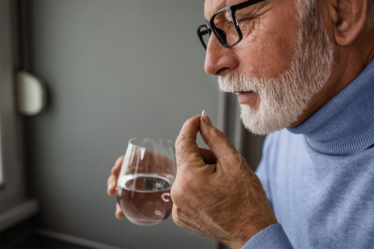 An older man holds a medication tablet in one hand and a glass of water in the other.